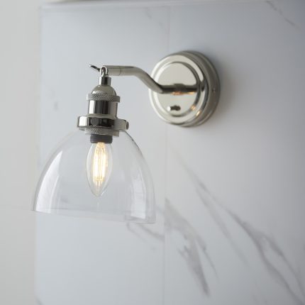 Close-up, slightly right-angle view of the Ferri Wall Light in Polished Nickel with a clear glass shade, installed on a marble effect wall in the kitchen.