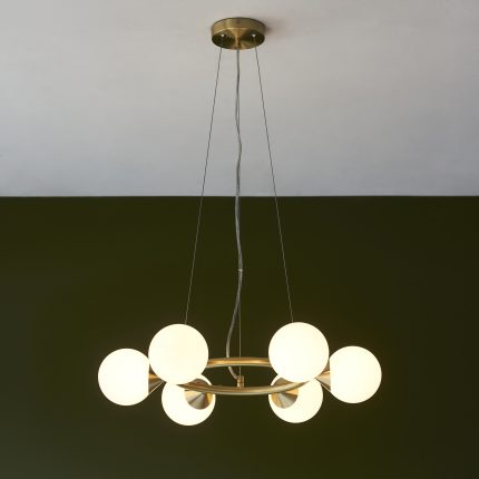 Nexus Chandelier with six round glass shades and a brass frame, hanging in a room with a green wall in the background.