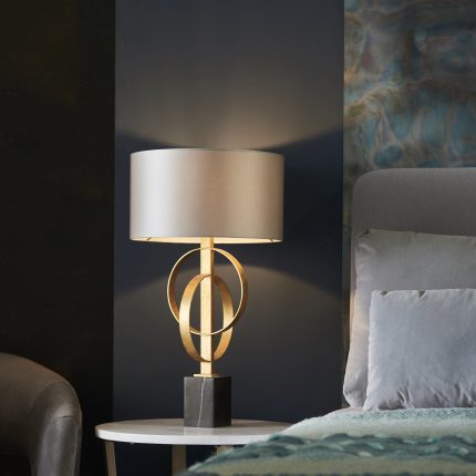 Close up of Oratio Table Lamp with Gold Leaf Hoops and Mink Drum Fabric Shade on bedside table next to a bed.