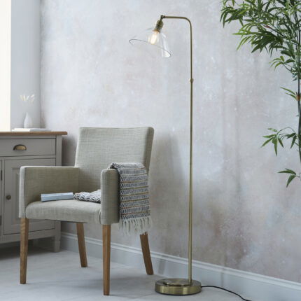 Freddo floor lamp with antique brass finish and clear glass shade positioned between reading chair and window.