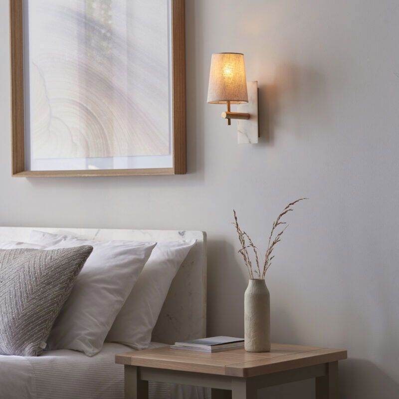 Wall light with brass arm and linen shade mounted above wooden bedside table next to bed with white bedding.