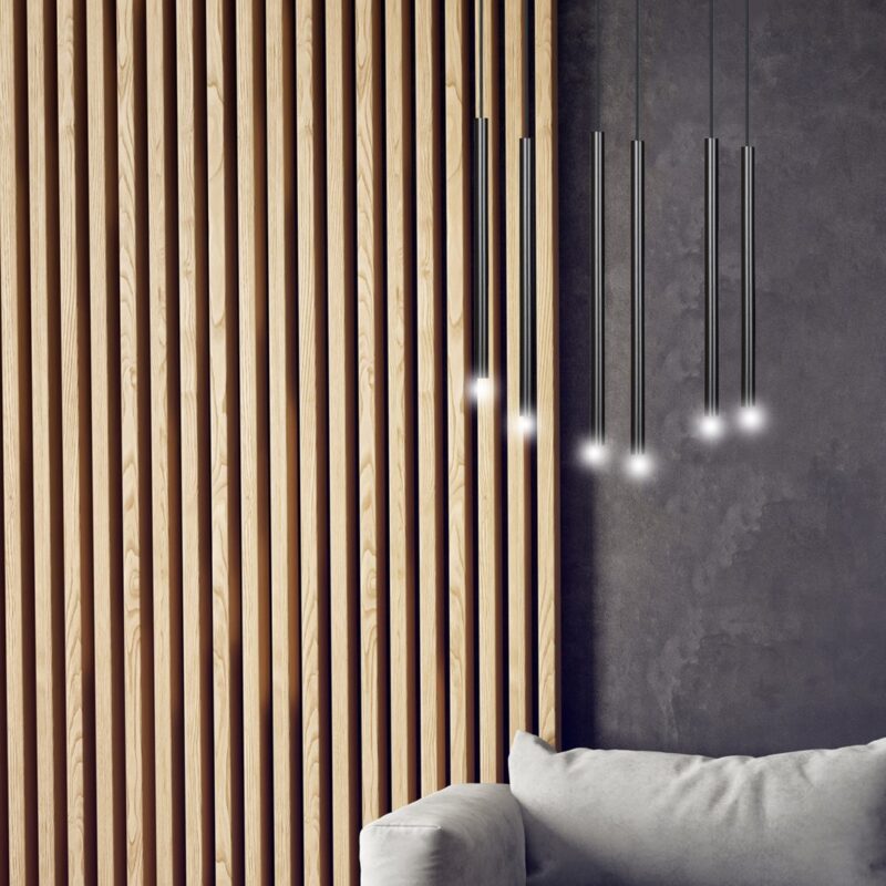 View of a lit six light pendant lamp hanging in a living room against wooden slat wall and above a sofa chair.