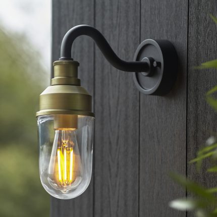 Side detail of the gold Navel Arched Arm Outdoor Wall Light, highlighting the clear glass shade encasing a glowing filament light bulb.