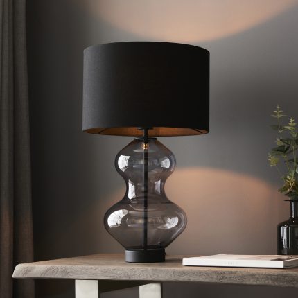 Close-up, slightly upward angle view of the right side of the Zephyr Matt Black Glass Touch Table Lamp, positioned on a light wooden table in a room, highlighting the intricate details of its black shade and grey glass base.