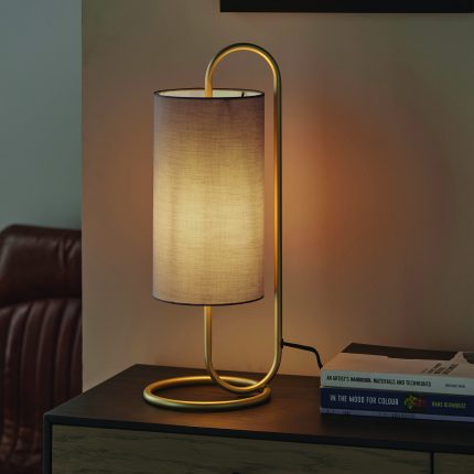 Eye level view of the Nostro Table Lamp with its light bulb turned on, featuring an oval antique brass base and a grey shade.