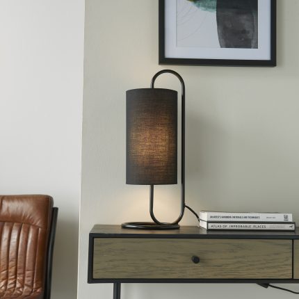 Eye level view of the Nostro Table Lamp with the bulb switched on, showing its matt black oval base and black cylinder shade, positioned on a wooden desk.